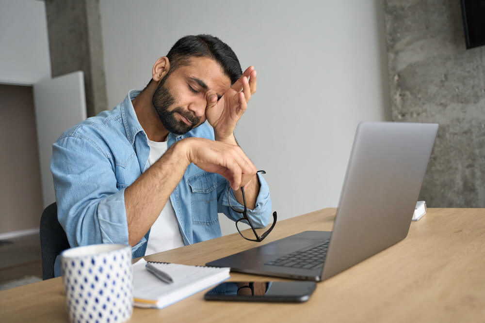 Man in front of laptop with his glasses in his hand, rubbing his eyes showing how tiring it is managing a tired workforce